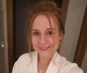 A woman with light brown hair is smiling at the camera while wearing a white robe and silver earrings, standing indoors.