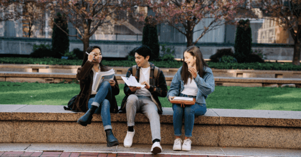 Three students sit on a stone bench outdoors, holding papers and notebooks, engaged in conversation and study, with trees and buildings in the background.