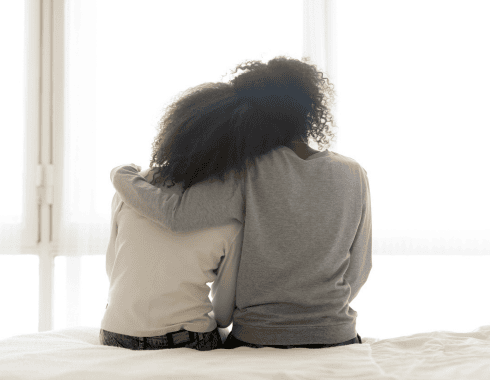 Two people with curly hair sit on a bed with their backs to the camera, one person’s arm around the other in a comforting gesture.