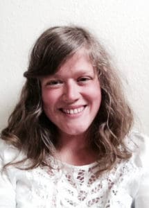 A person with wavy, light brown hair and a white lace top smiles at the camera in front of a plain light-colored wall.