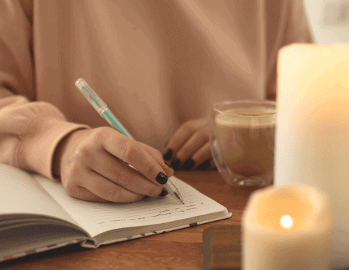 A person writes in a notebook with a pen while sitting at a table with a cup of coffee and lit candles nearby.