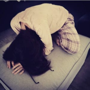 A person in pajamas is kneeling on a cushion, bent forward with their head down and hair covering their face. Glasses are lying next to their hand.