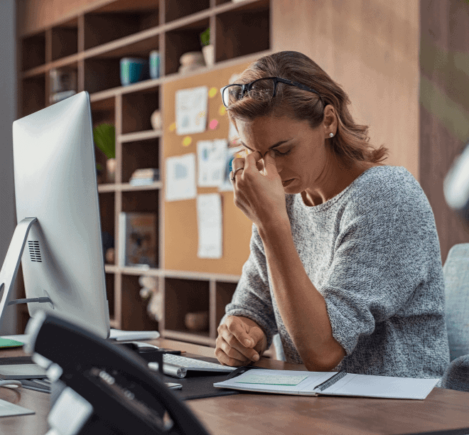 A woman sits at a desk in an office, holding her head and looking stressed while working on paperwork and using a computer.