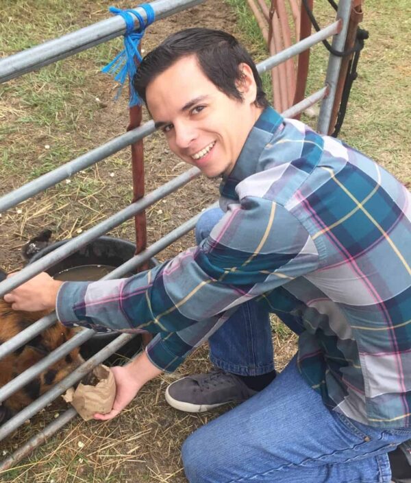 A man in a plaid shirt crouches by a metal fence, smiling while feeding an animal through the bars.