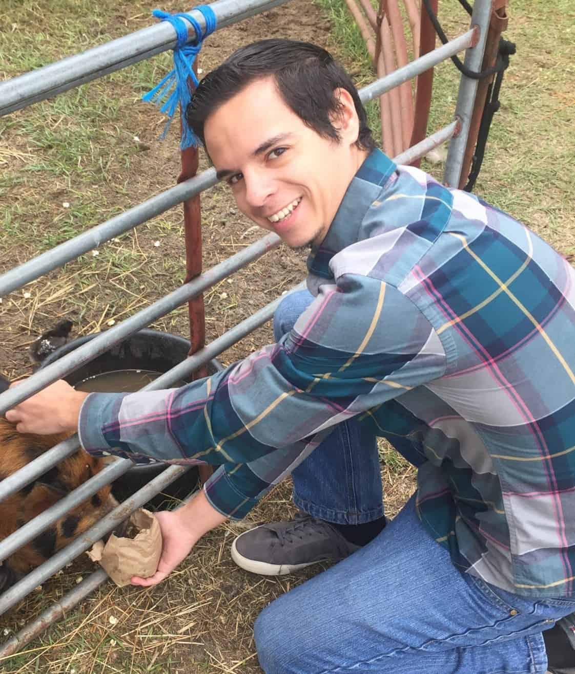 A man in a plaid shirt crouches by a metal fence, smiling while feeding an animal through the bars.