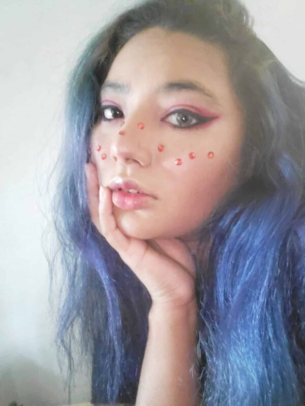 Person with long blue hair and pink makeup, including pink eyeliner and decorative pink dots on cheeks and nose, resting their face on their hand.
