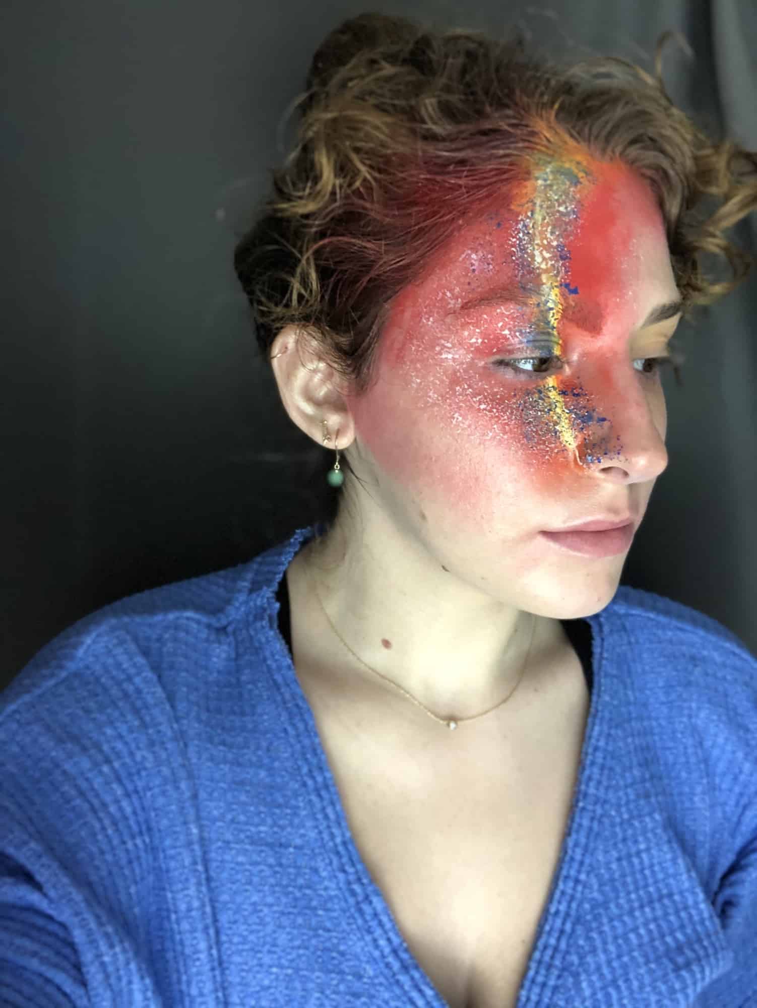 A person with curly hair wears bold, colorful makeup covering half their face, featuring red, orange, and glitter accents. They are dressed in a blue top against a dark background.