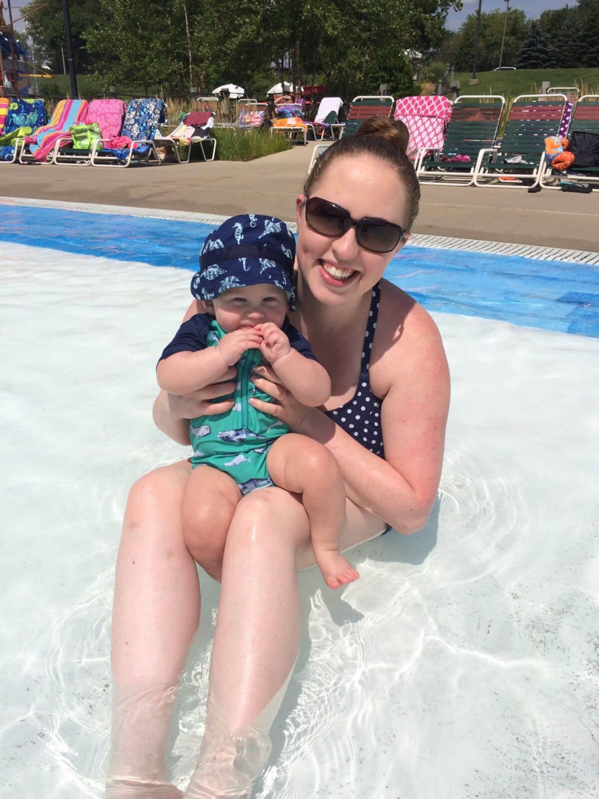 Woman wearing sunglasses sits in a shallow pool holding a baby in a sunhat and swimsuit; colorful towels and chairs are visible in the background.