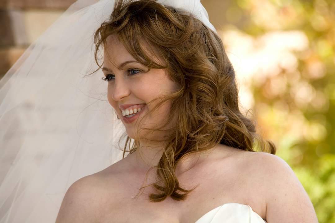 A woman in a strapless white dress and veil smiles outdoors, with blurred greenery and sunlight in the background.