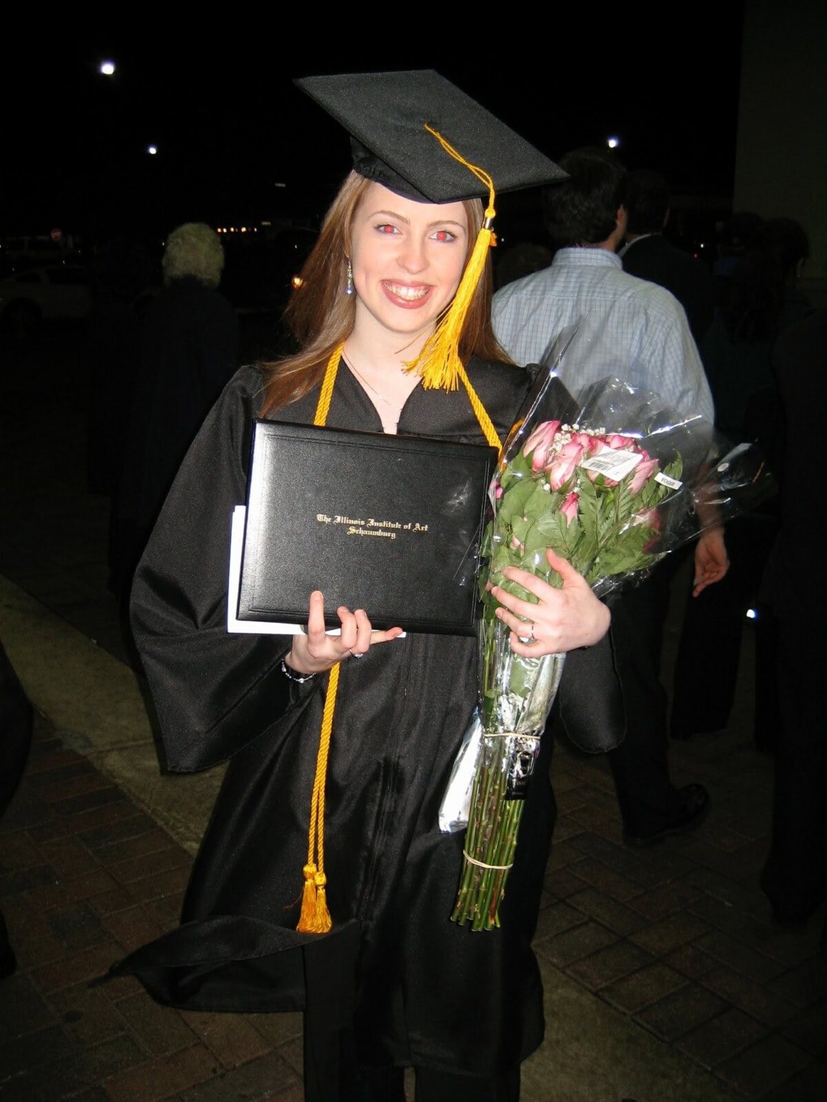 A graduate in a cap and gown holds a diploma and a bouquet of flowers, smiling at the camera during an outdoor graduation ceremony at night.