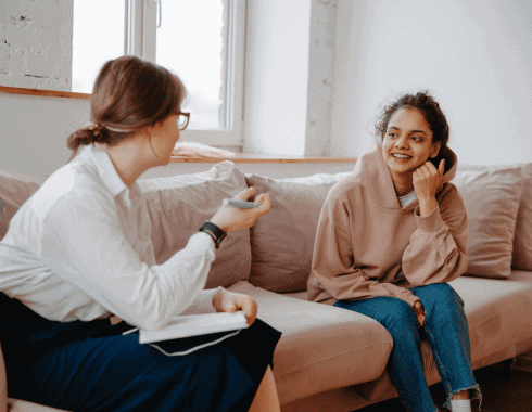 Two women are sitting on a couch facing each other; one is holding a notebook and pen, while the other is smiling and listening attentively.