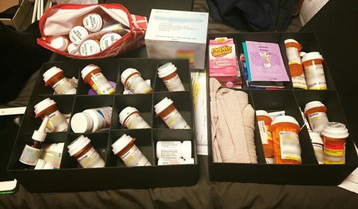 Two black boxes hold numerous prescription pill bottles, over-the-counter medications, Pepto-Bismol, and packets, along with a few personal items on a bed.
