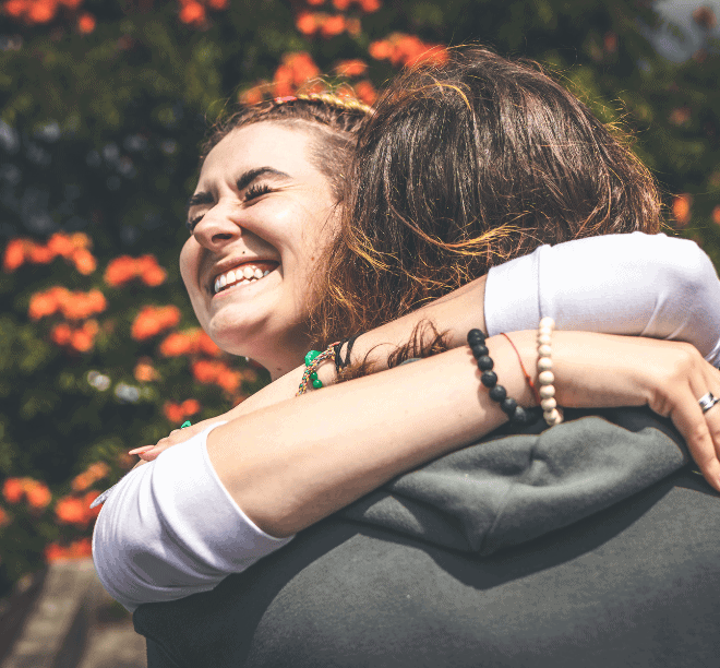 Two people embrace outdoors, one smiling broadly with eyes closed. Bright orange flowers and greenery are visible in the background.
