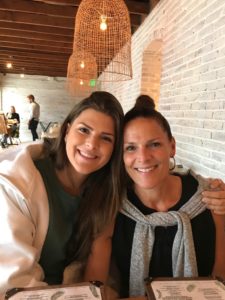 Two women sit closely together at a restaurant table, smiling at the camera. Menus are visible in front of them and the background features white brick walls and hanging lights.