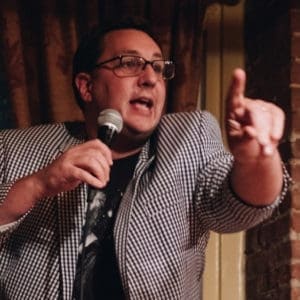 A man wearing glasses and a checkered jacket speaks into a microphone and points with his finger, standing indoors near a brick wall.