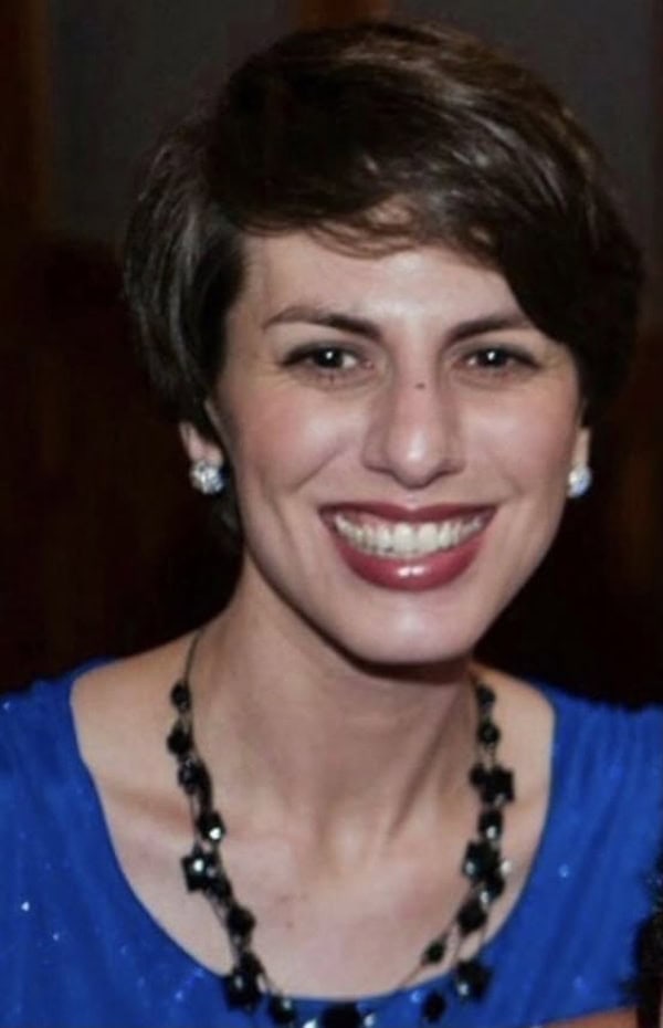 A woman with short brown hair, wearing a blue top, stud earrings, and a black beaded necklace, smiles at the camera.