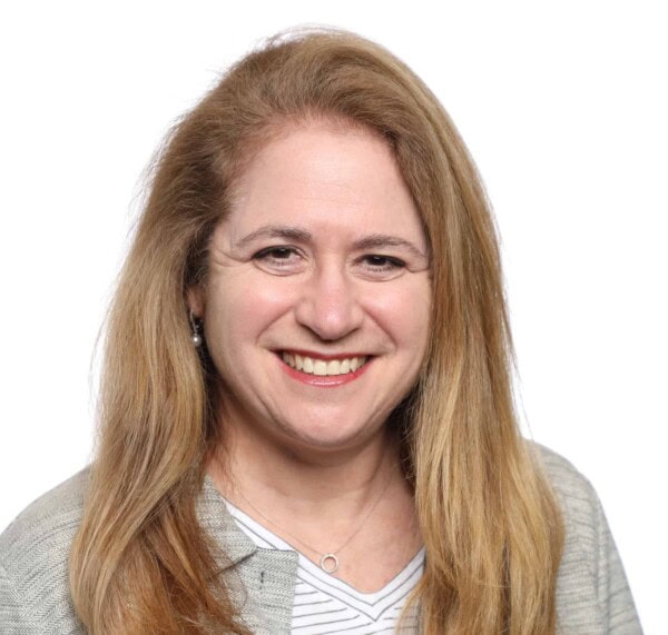 A woman with long blonde hair, wearing a gray cardigan and striped top, smiles at the camera against a white background.
