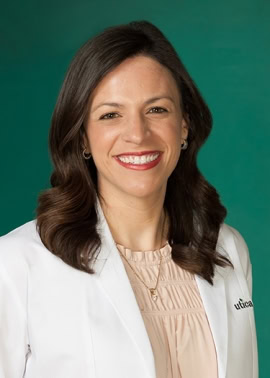 Woman with brown hair wearing a white coat and light pink blouse, smiling in front of a green background.