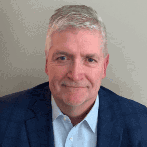 A middle-aged man with short gray hair, wearing a blue suit jacket and light blue shirt, sits in front of a plain background and smiles slightly at the camera.