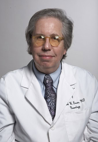 A middle-aged person wearing glasses, a white medical coat, and a patterned tie poses against a plain background. The coat has embroidered text that includes 