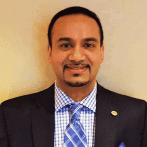 A man in a suit and checked shirt with a blue patterned tie, standing against a plain light-colored background, smiling at the camera.