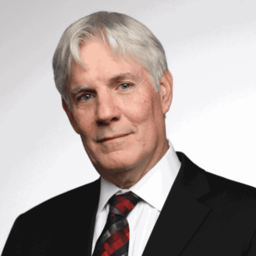 A man with short gray hair wearing a black suit, white shirt, and patterned tie, posed against a plain light background.