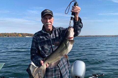 An older man in a plaid shirt and cap stands on a boat holding a large fish with a grip tool over a lake.