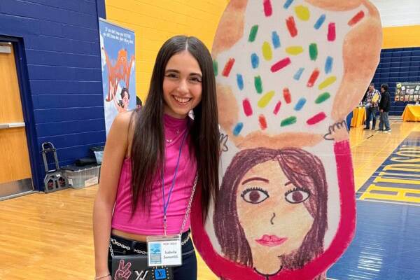 A girl in a pink top stands next to a large drawing of a person holding a sprinkle-covered donut at an indoor event.