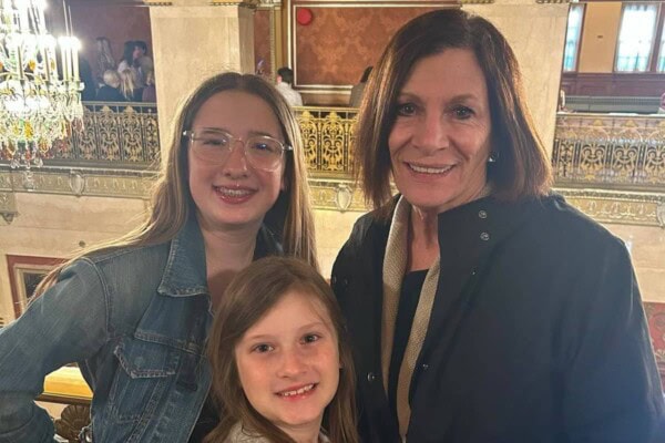 Three people, two children and one adult, stand together indoors in front of ornate railings and a chandelier, smiling at the camera.