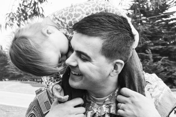 A man in military uniform smiles while a young child sits on his shoulders, hugging his head. Trees are visible in the background.