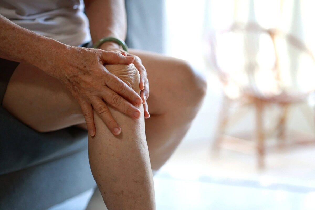 Close-up of a person sitting and holding their knee with both hands, suggesting discomfort or pain in the knee.