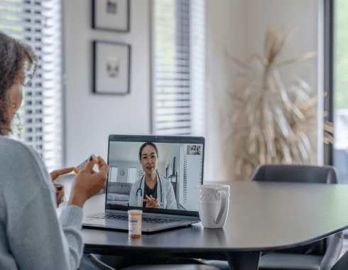 A person holds a pill bottle while having a video call with a healthcare professional on a laptop at a table with a mug and another pill bottle.
