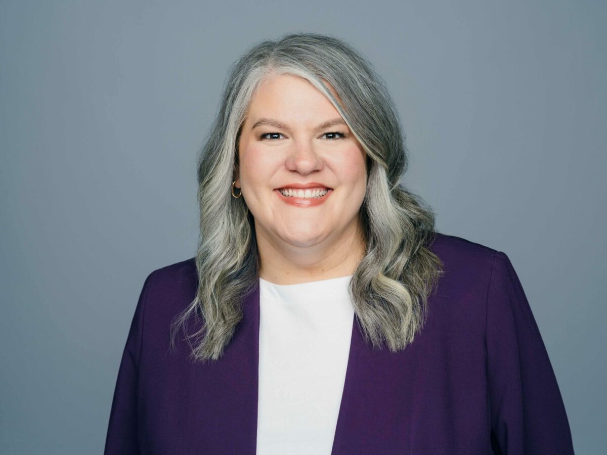 Smiling woman with long gray hair, wearing a white top and purple blazer, posing in front of a plain gray background.