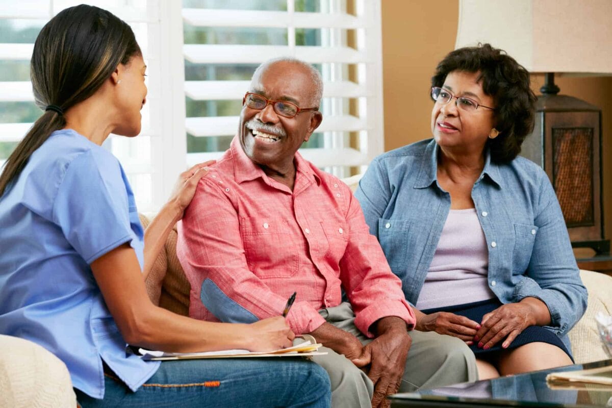 A healthcare professional talks with an older man and woman, taking notes as they sit together in a living room.