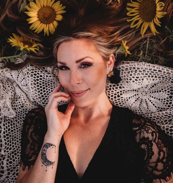 Woman with a crescent moon tattoo on her wrist lies on a crocheted blanket surrounded by sunflowers, touching her face and looking at the camera.