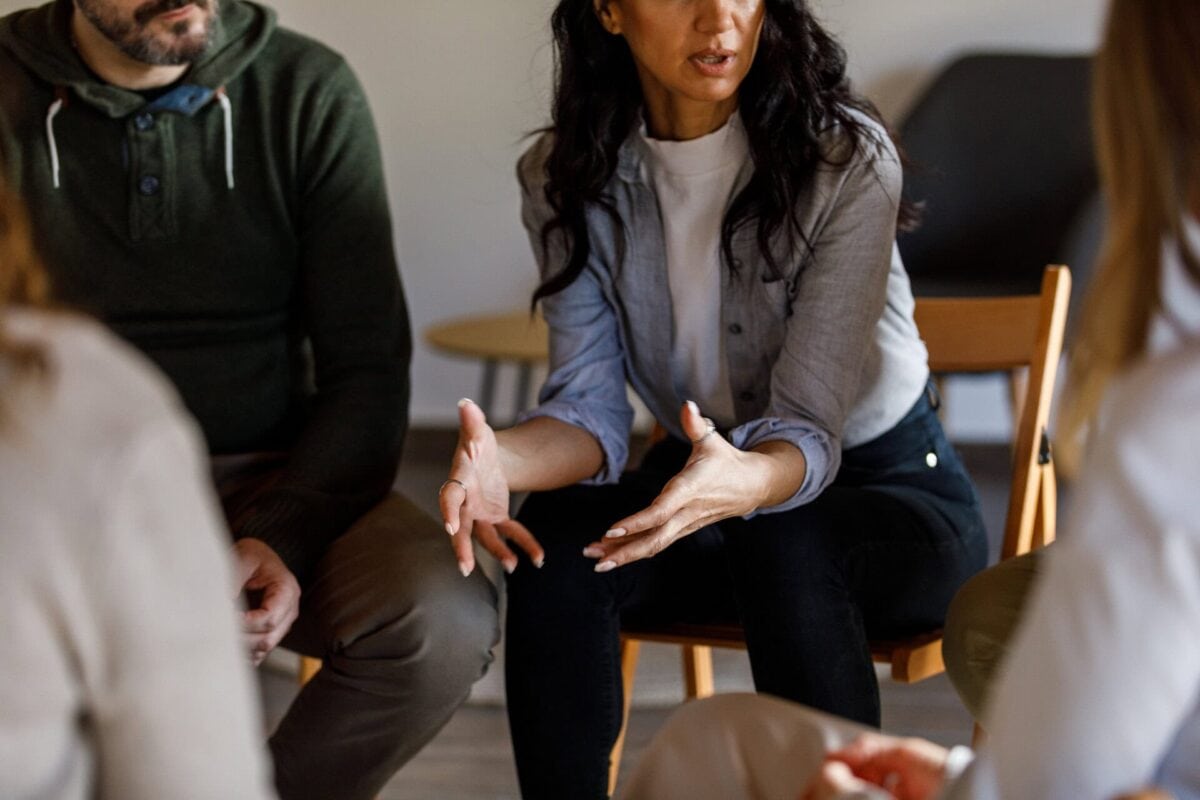A woman sits in a chair, gesturing with her hands while speaking to others in a small group discussion.