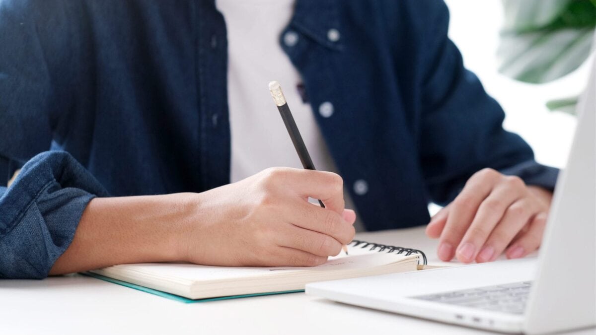 A person sits at a desk writing in a notebook with a pencil, with an open laptop in front of them.