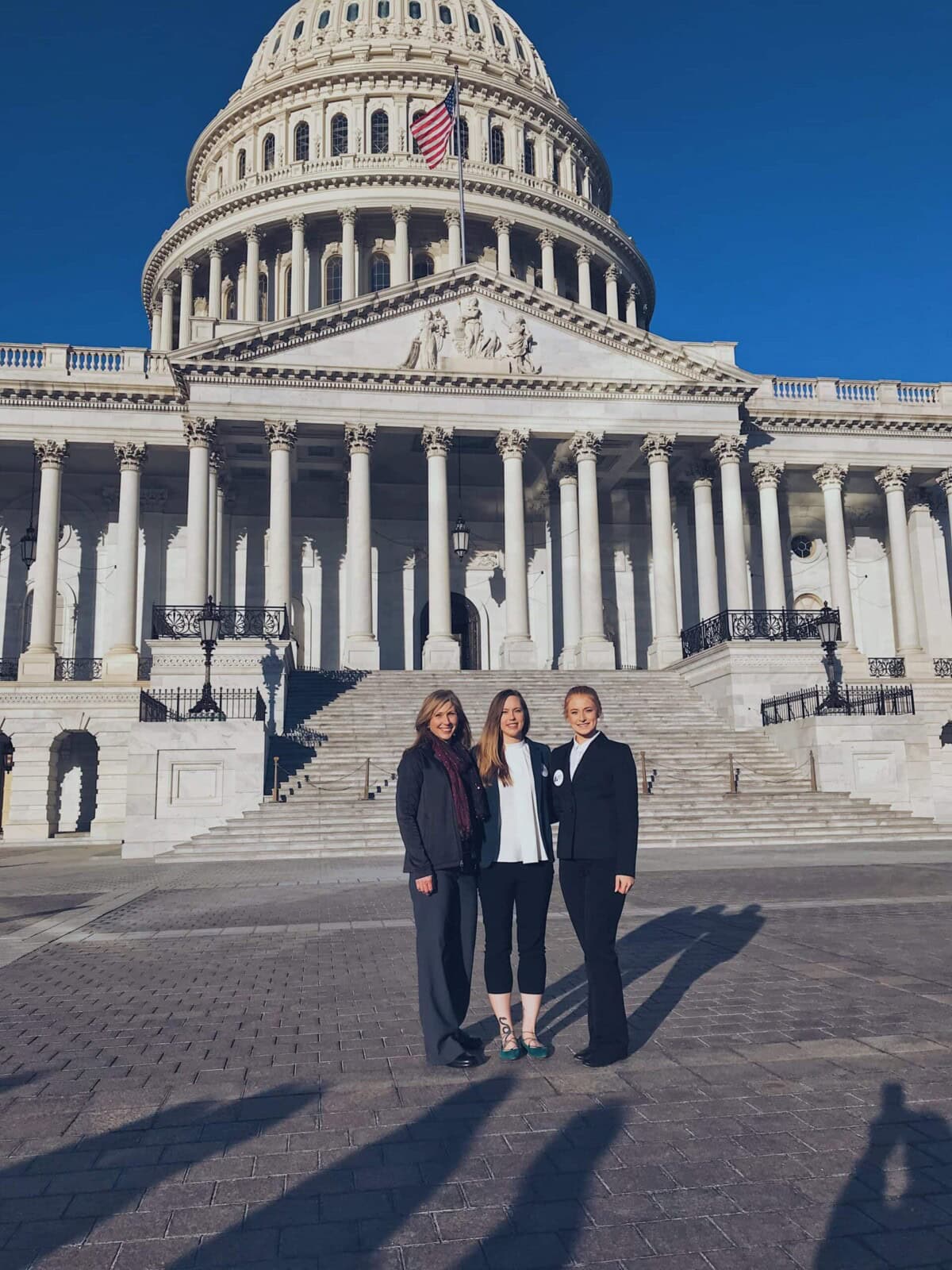 Three women stand in front of the U.S. Capitol building on a sunny day.