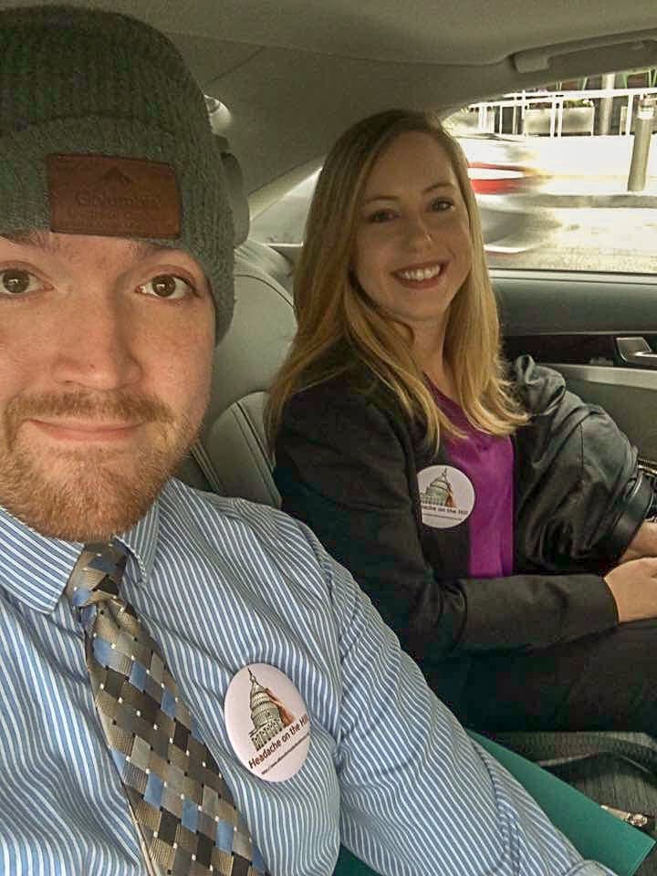 Two people sitting in a car, both wearing buttons with a Capitol building graphic and the text "Hearing on the Hill." The man is wearing a beanie, tie, and striped shirt.