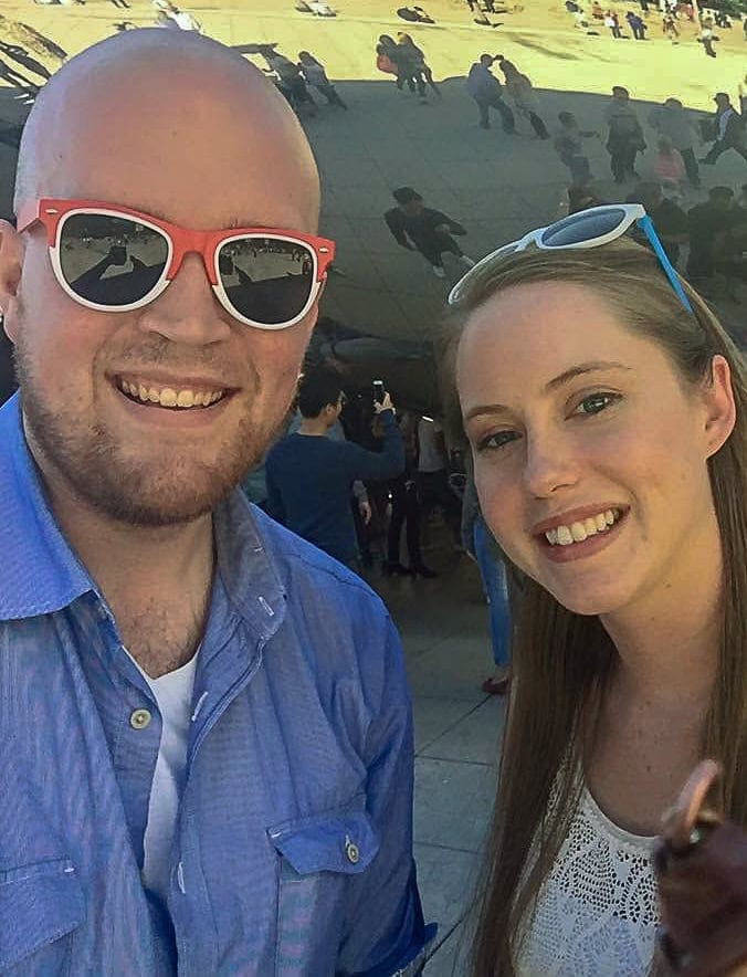 A man and a woman wearing sunglasses smile for a selfie outdoors with a reflective sculpture and other people in the background.