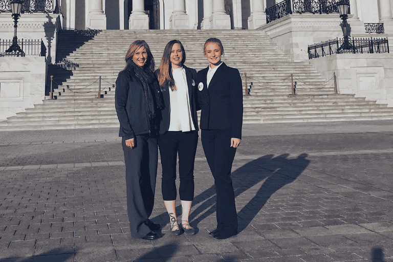 Three women stand smiling in front of a large stone staircase on a sunny day, dressed in business and semi-formal attire.