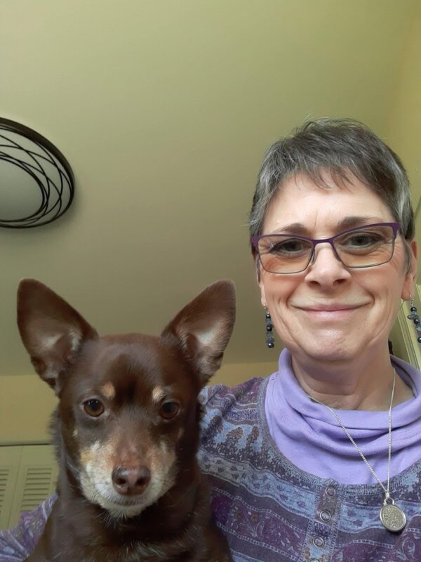 A person with short gray hair and glasses smiles for a selfie with a small brown dog indoors.