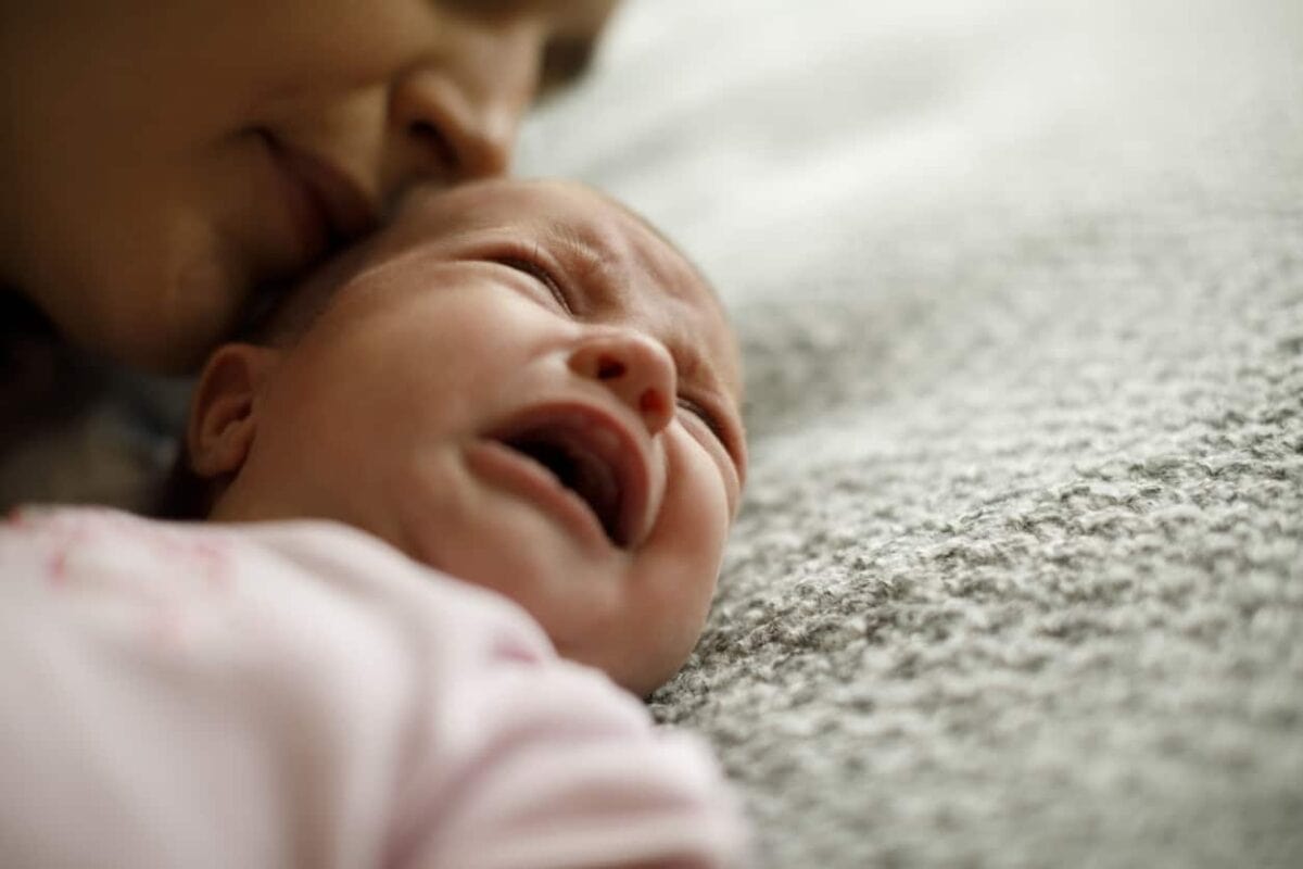 A close-up of a crying baby lying on a soft surface, with an adult's face partially visible, comforting the baby.