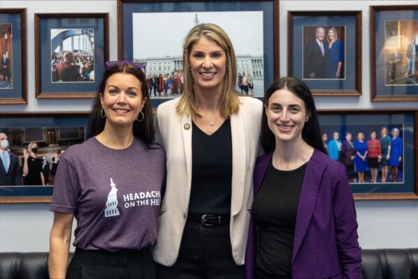 Three women stand smiling in front of a wall with framed photos; one woman wears a 