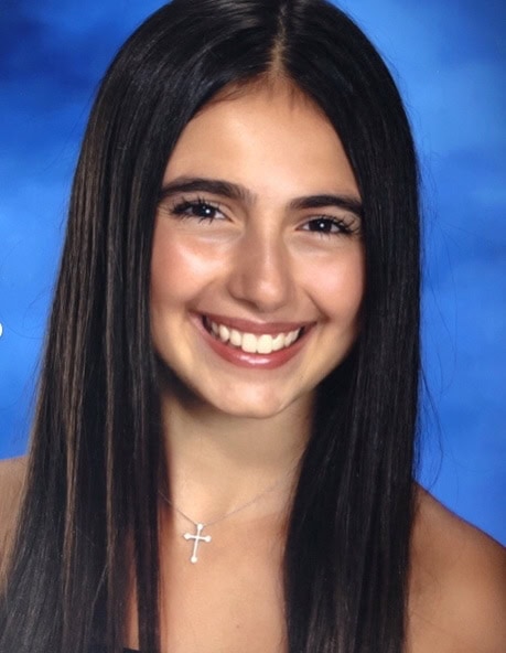 A young woman with long, straight dark hair smiles in front of a blue background, wearing a necklace with a cross pendant.