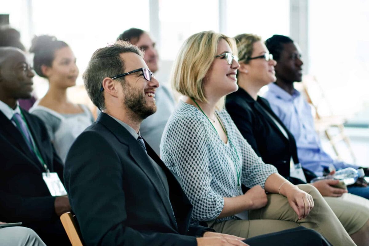 A group of people sit in rows, attentively listening and smiling at an indoor event or presentation.