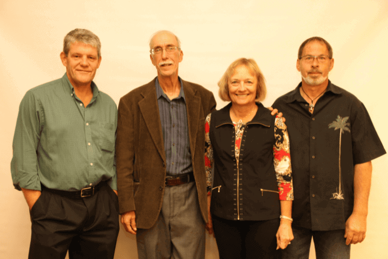 Four adults stand side by side in front of a plain light background, posing for a group photo and looking at the camera.