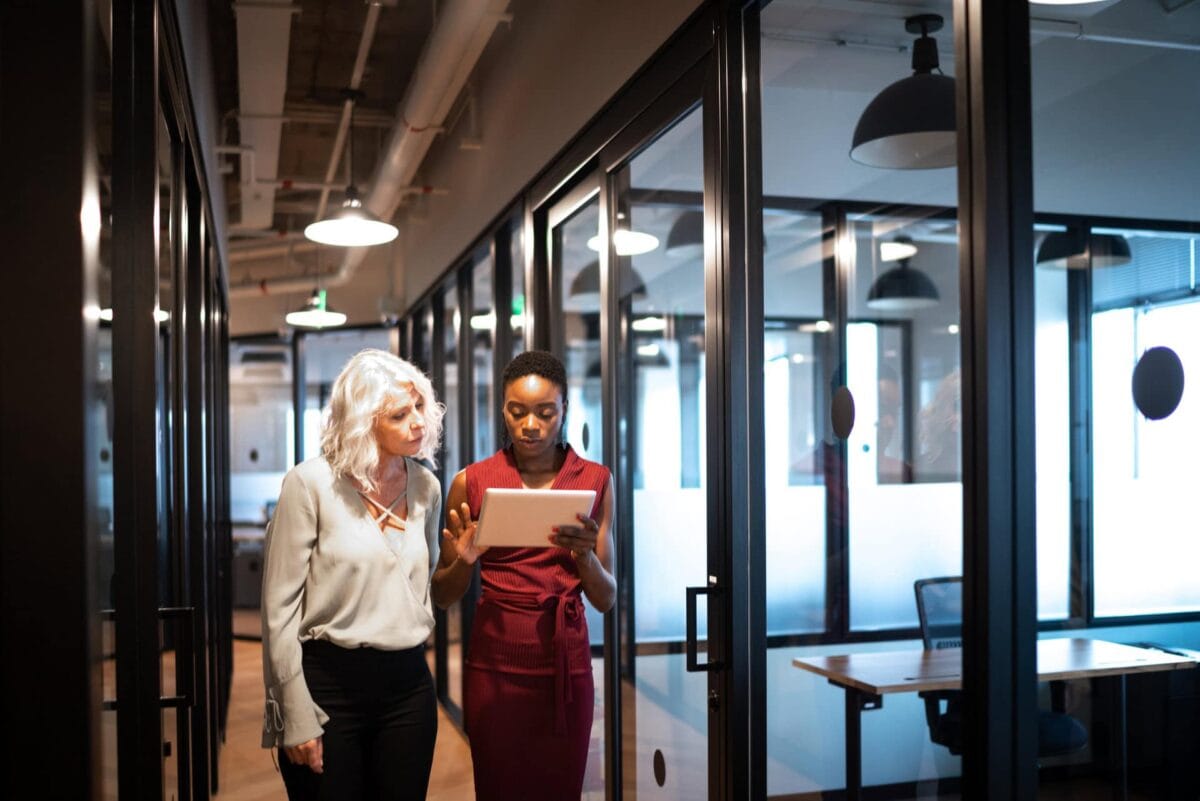 Two women walk through an office hallway while looking at a digital tablet together.