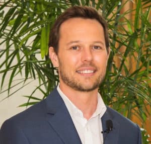 A man with short brown hair and a beard, wearing a blue suit and white shirt, stands in front of green plants. He has a small microphone attached to his jacket.