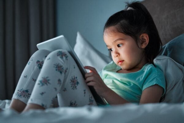 A young girl in pajamas sits on a bed using a tablet, with soft lighting illuminating her face.
